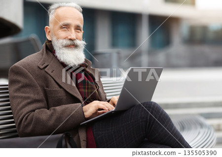 Wealthy grey-haired aged man with long beard in warm stylish outfit sitting on the bench by office building, using laptop, working online, chatting with clients or websurfing, empty space Wealthy grey-haired aged man with long beard in warm stylish outfit sitting on the bench by office building, using laptop, working online, chatting with clients or websurfing, empty space 135505390