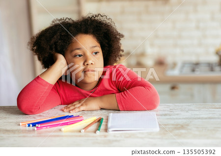 Boredom. Cute little bored black girl sitting at table with notebook and pencils in kitchen, thoughtful african american female child resting head on hand and looking away, feeling lonely, closeup 135505392