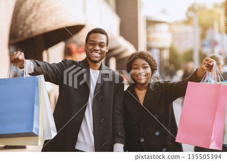 A lot of shopping in seasonal sale and Black Friday before holidays. Happy young african american male and female in coats showing many colorful shopping bags and express success near town shop A lot of shopping in seasonal sale and Black Friday before holidays. Happy young african american male and female in coats showing many colorful shopping bags and express success near town shop 135505394