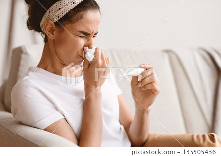 Closeup ofs ick young woman sitting on couch and sneezing, measuring body temperature, home interior. Brunette lady got cold or flu, having fever and runny nose, copy space. Cold, flue, coronavirus Closeup ofs ick young woman sitting on couch and sneezing, measuring body temperature, home interior. Brunette lady got cold or flu, having fever and runny nose, copy space. Cold, flue, coronavirus 135505436