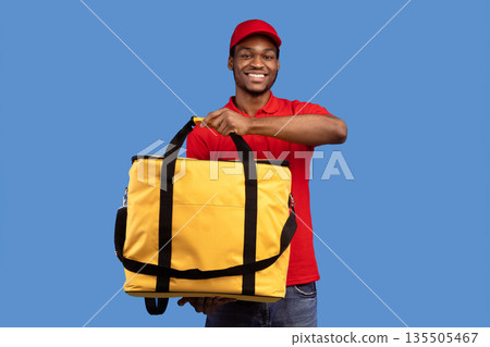 Fast Food Delivery Service Cocnept. Portrait of smiling black male courier wearing red uniform holding yellow thermo backpack bag and showing it to camera isolated on blue studio background Fast Food Delivery Service Cocnept. Portrait of smiling black male courier wearing red uniform holding yellow thermo backpack bag and showing it to camera isolated on blue studio background 135505467