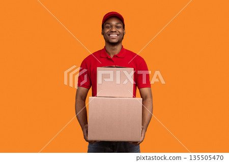 Delivery Service Concept. Smiling african american male courier in red cap and uniform holding pile of cardboard boxes, carrying packages, standing isolated over orange studio background Delivery Service Concept. Smiling african american male courier in red cap and uniform holding pile of cardboard boxes, carrying packages, standing isolated over orange studio background 135505470