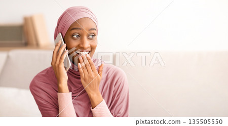 Modern communications concept. Surprised Muslim woman in hijab talking on cellphone at home, chatting online with friend. Lovely Arab lady in headscarf gossiping, discussing shocking news 135505550