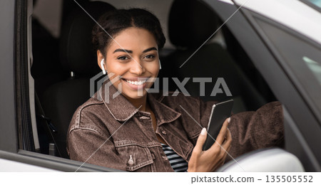 Driving work, modern taxi driver and profession in city. Smiling young african american woman in wireless headphones holding phone, sitting in silver car, looking through open window, copy space Driving work, modern taxi driver and profession in city. Smiling young african american woman in wireless headphones holding phone, sitting in silver car, looking through open window, copy space 135505552