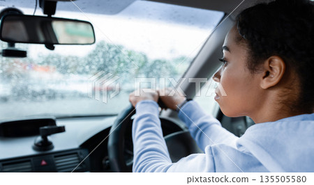 Working as taxi driver in city in bad rainy weather and way to job. Focused young african american lady drives car and holds steering wheel, looks at road on windshield with raindrops, empty space Working as taxi driver in city in bad rainy weather and way to job. Focused young african american lady drives car and holds steering wheel, looks at road on windshield with raindrops, empty space 135505580