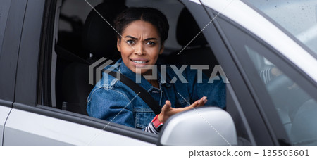 Conflict, difficulties and problems on city road. Excited angry millennial african american lady driver holding steering wheel, pointing at camera and looking out window and swears in car, side view Conflict, difficulties and problems on city road. Excited angry millennial african american lady driver holding steering wheel, pointing at camera and looking out window and swears in car, side view 135505601