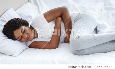 Young black woman touching her belly, laying in bed, side view, copy space. Sad african american lady suffering from abdominal pain, hugging her tummy and crying, home interior Young black woman touching her belly, laying in bed, side view, copy space. Sad african american lady suffering from abdominal pain, hugging her tummy and crying, home interior 135505648