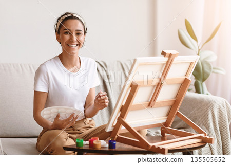 Happy young woman with easel and palette painting at home, copy space. Beautiful lady painting for hobby in living room, practicing with paint, brushes, easel and colors. Hobby, creativity concept Happy young woman with easel and palette painting at home, copy space. Beautiful lady painting for hobby in living room, practicing with paint, brushes, easel and colors. Hobby, creativity concept 135505652