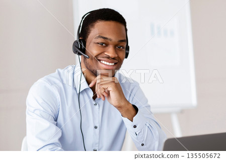 Cheerful black guy worker call center representative with headset using laptop, smiling at camera, african american man having business training or conference online in office, copy space 135505672
