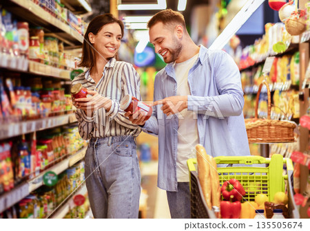 Millennial Family Couple Shopping Groceries In Supermarket Buying Food Walking With Trolley In Grocery Shop Indoor. Cheerful Customers Buy Supply Essentials Standing Near Shelves In Store Aisle Millennial Family Couple Shopping Groceries In Supermarket Buying Food Walking With Trolley In Grocery Shop Indoor. Cheerful Customers Buy Supply Essentials Standing Near Shelves In Store Aisle 135505674
