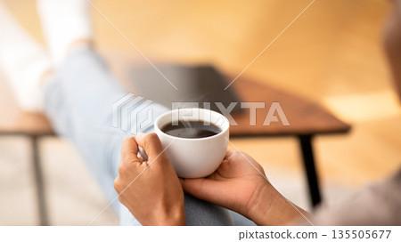 Closeup of young black woman warming her hands on cup of coffee, relaxing at home, empty space. Unrecognizable lady with aromatic hot beverage enjoying peaceful relaxing time on day off 135505677