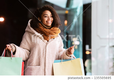 Portrait of smiling african american woman looking at fashion clothes in shop window of the mall, walking in the evening near shopping center. Winter Gifts, Sales And Discount, Retail, Purchase 135505695