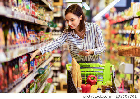Portrait Of Millennial Lady Holding And Using Smartphone Buying Food Groceries Walking In Supermarket With Trolley Cart. Female Customer Shopping With Checklist, Taking Products From Shelf At The Shop 135505701