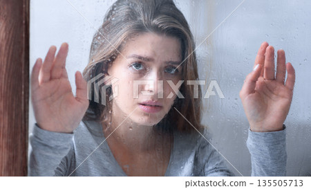 Beautiful millennial woman feeling sad and lonely because of bad weather, touching window glass with rain drops. Young lady feeling depressed, having seasonal affective disorder 135505713