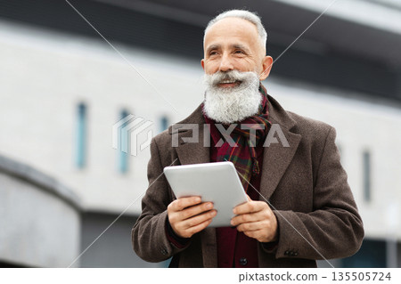 Cheerful aged man with long grey beard holding digital tablet while walking by street, looking at copy space and smiling. Senior businessman in stylish outfit with pad standing by office building 135505724