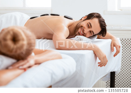 Couples Hot Stone Massage. Contented Husband Smiling To Wife Lying And Relaxing On Massage Therapy At Spa Salon During Romantic Honeymoon. Beauty, Relaxation And Wellness. Selective Focus 135505728