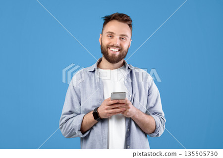 Portrait of cool bearded guy using mobile phone on blue studio background. Cheerful young man with modern gadget browsing internet, checking email, posting in social media 135505730