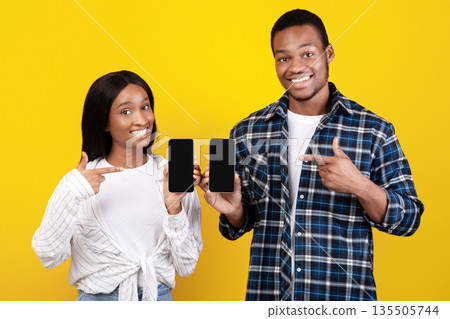 Online app for purchases and messages from stores. Smiling zoomers african american couple showing smartphones with blank screens and pointing fingers at gadgets, isolated on yellow background 135505744