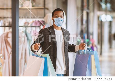Time to buy goods at low price during pandemic covid-19 and social distance. Happy young african american male in protective masks holding many colored shopping bags in city mall, free space 135505754