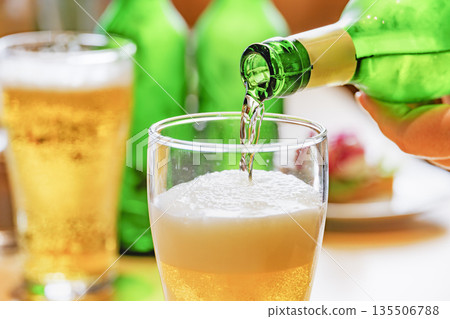Golden beer poured from a green bottle into a glass with foam Golden beer poured from a green bottle into a glass with foam 135506788