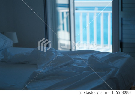 Cozy bedroom with rumpled white sheets and open balcony overlooking serene ocean view 135508995