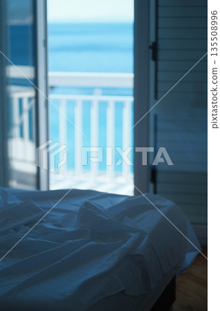 Cozy bedroom with rumpled white sheets and ocean view through open balcony doors 135508996