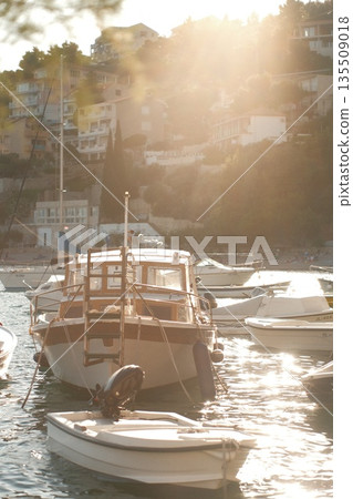 Boats anchored in tranquil harbor with sunlit reflections and scenic coastal backdrop Boats anchored in tranquil harbor with sunlit reflections and scenic coastal backdrop 135509018