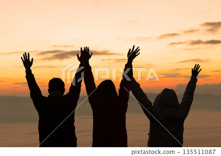 Silhouettes of friends raising hands on a mountain at sunrise above a sea of fog, dramatic sky behind. Concept of teamwork, unity, celebration, freedom, hope, success and new beginnings. 135511087