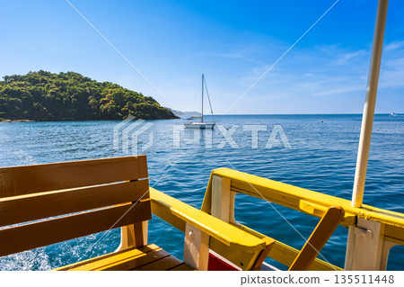 View from a ferry deck of a white sailboat anchored in the calm blue bay of Koh Samed, Rayong, Thailand View from a ferry deck of a white sailboat anchored in the calm blue bay of Koh Samed, Rayong, Thailand 135511448