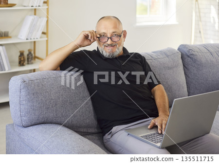 Mature man with laptop on comfy sofa at home, smiling and lifting reading glasses 135511715