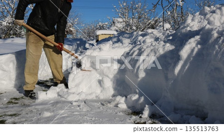 The lower part of a man holding a wooden shovel and clearing a large amount of snow on a sunny day, snow removal from a residential area in a rural area 135513763