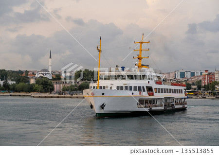 A ferry navigates the Bosphorus Strait, gliding past the vibrant city skyline, historic structures, and lush greenery under a moody sky. A ferry navigates the Bosphorus Strait, gliding past the vibrant city skyline, historic structures, and lush greenery under a moody sky. 135513855