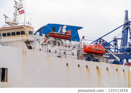 A cargo ship is moored in the Bosphorus, featuring bright orange rescue boats and industrial cranes in the background under a cloudy sky. A cargo ship is moored in the Bosphorus, featuring bright orange rescue boats and industrial cranes in the background under a cloudy sky. 135513856