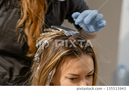 A stylist applies hair dye using foil highlights while a client relaxes in a salon chair during the afternoon. 135513943