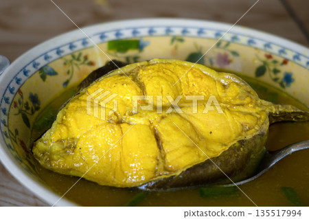 Close-up of ikan sembilang asem asem or Indonesian sour soup of Eel-tailed Catfish dish in a floral bowl with a spoon, on a wooden table with warm tones. Close-up of ikan sembilang asem asem or Indonesian sour soup of Eel-tailed Catfish dish in a floral bowl with a spoon, on a wooden table with warm tones. 135517994