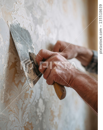 Skilled Worker's Hands Using a Wide Metal Spatula to Scrape and Remove Old, Patterned Wallpaper from a Wall During Home Renovation, Repair, DIY Decorating Projects, Illustrating the Preparation Stage 135518639