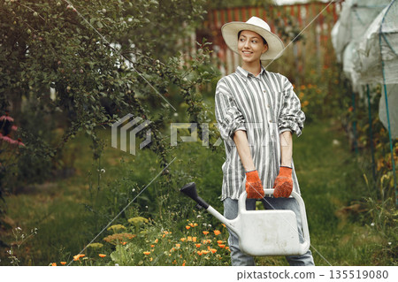 Gardening in summer. Woman watering flowers with a watering can. Girl wearing a hat. 135519080