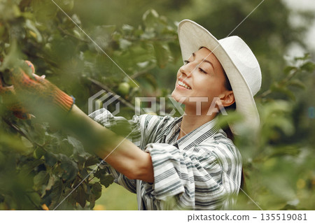 Young woman taking care of plants. Brunette in a hat and gloves. 135519081