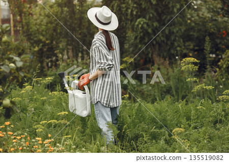 Gardening in summer. Woman watering flowers with a watering can. Girl wearing a hat. 135519082