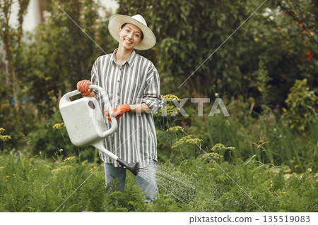 Gardening in summer. Woman watering flowers with a watering can. Girl wearing a hat. 135519083