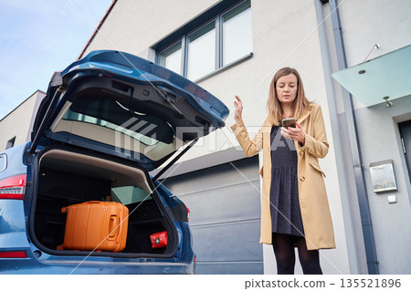 Woman standing near car with open trunk and suitcase, holding phone and raising hand Woman standing near car with open trunk and suitcase, holding phone and raising hand 135521896