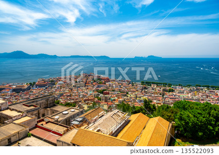 Panoramic top view of Naples rooftops from Castel Sant Elmo, with the sea in the distance and Mount Vesuvius rising above the city on a clear summer day. 135522093