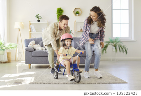 Little girl rides tricycle at home as supportive parents cheer and smile 135522425