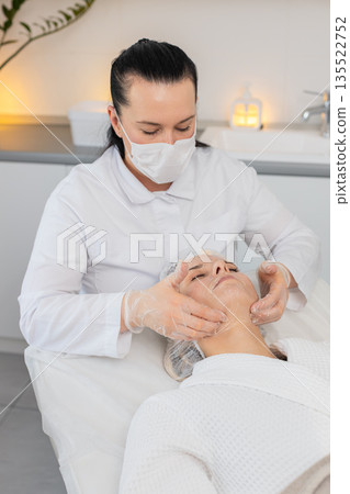 Beautician cosmetologist massaging peeling serum on middle aged young adult woman face in clinic Beautician cosmetologist massaging peeling serum on middle aged young adult woman face in clinic 135522752