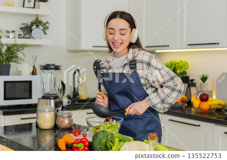Young woman having fun with utensils while tossing keto veggie salad at home indoors kitchen modern Young woman having fun with utensils while tossing keto veggie salad at home indoors kitchen modern 135522753
