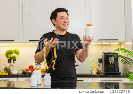Asian man in kitchen holding supplement bottle with tape measure celebrating fit healthy weight loss 135522758