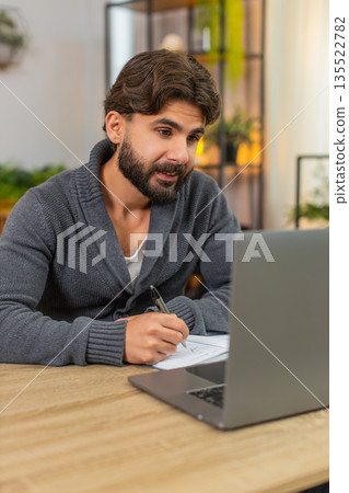 Indian man studies online on laptop notes from ebook to notebook focused learning calm mood session Indian man studies online on laptop notes from ebook to notebook focused learning calm mood session 135522782