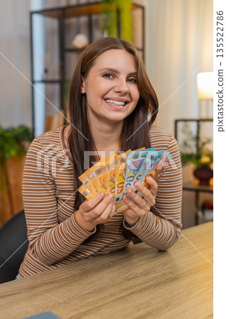 Young woman counts cash euro bills at table smiles pleased about salary lottery win savings income Young woman counts cash euro bills at table smiles pleased about salary lottery win savings income 135522786