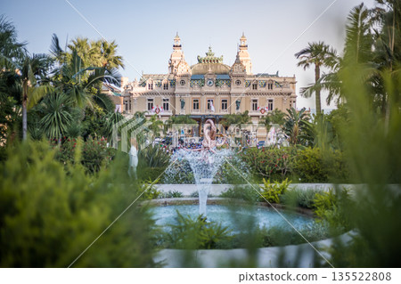Elegant fountain surrounded by palm gardens on the square in front of the Monte Carlo Casino, Monaco. Iconic luxury landmark with historic architecture under clear blue sky. Elegant fountain surrounded by palm gardens on the square in front of the Monte Carlo Casino, Monaco. Iconic luxury landmark with historic architecture under clear blue sky. 135522808