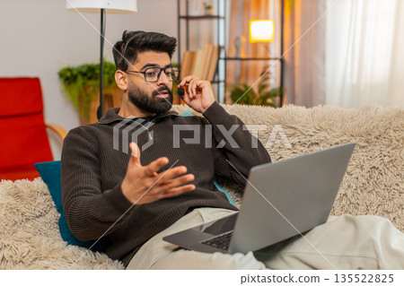 Indian man using wireless headset speaks on laptop video call with client while working at home 135522825
